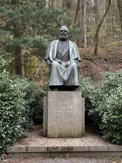 Eine Bronzestatue von Karl Marx, sitzend, in einem bewaldeten Gebiet gelegen. Die Statue steht auf einem Steinsockel mit der Inschrift seines Namens. Umgeben von Grün und Bäumen, ist die Umgebung ruhig und natürlich.