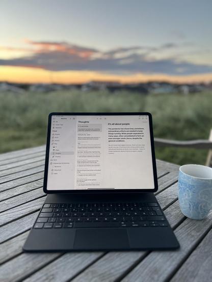 Laptop open on a wooden table displaying a text document titled "Thoughts," with a blurred background capturing a sunset sky and a grassy field. A ceramic mug sits to the right of the laptop.