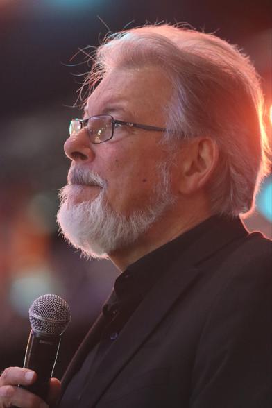 A man with gray hair and a beard, wearing glasses and a black shirt, is holding a microphone and looking thoughtfully into the distance. Soft lighting highlights his profile.