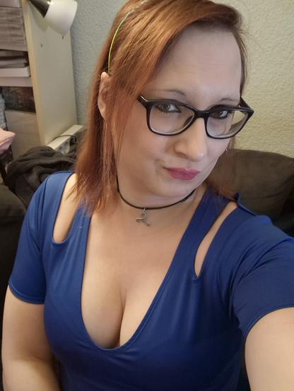 Das Bild zeigt eine Frau mit roten Haaren, die eine Brille trägt. Sie trägt eine blaue Top mit einem tiefen Ausschnitt und Schlitzen an den Schultern. Um ihren Hals hat sie eine schwarze Kette mit einem silbernen Anhänger. Im Hintergrund ist ein Teil eines Sofas und ein Lampenständer zu sehen, was auf eine Wohnraumumgebung hinweist. Die Frau lächelt leicht in die Kamera.