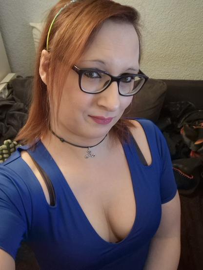 Das Bild zeigt eine Frau mit roten Haaren, die eine Brille mit schwarzen Rahmen trägt. Sie hat ein blaues Top mit einem tiefen V-Ausschnitt und zwei offene Schulterpartien, die einen Teil ihrer Schultern zeigen. Um ihren Hals trägt sie eine schwarze Kette mit einem silbernen Anhänger. Ihre Haare sind mit einem grünen Haarband zusammengehalten. Im Hintergrund ist ein dunkelgrauer Sofa und einige Kleidungsstücke zu sehen. Die Frau lächelt leicht in die Kamera.