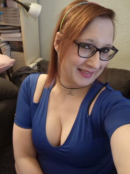 Das Bild zeigt eine Frau mit roten Haaren, die ein blaues Top mit einem tiefen Ausschnitt und offenen Schultern trägt. Sie trägt eine schwarze Kette mit einem kleinen Anhänger und eine schwarze Haarspange mit bunten Details. Sie lächelt in die Kamera und trägt eine Brille mit schwarzen Rahmen. Im Hintergrund ist ein Teil eines Sofas und ein Regal mit Büchern und einem Lampenschirm zu sehen. Die Frau scheint in einem Wohnzimmer zu sein.