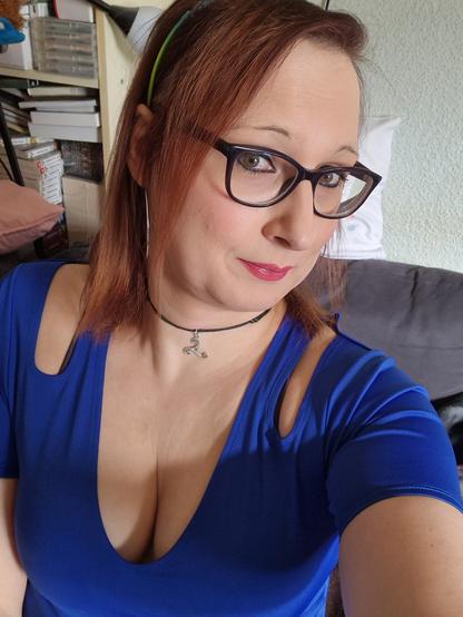 Das Bild zeigt eine Frau mit roten Haaren, die eine blaue Top mit einem tiefen Ausschnitt trägt. Sie hat eine schwarze Brille mit einem auffälligen Rahmen und ein silbernes Halsband mit einem Anhänger. Ihre Haare sind mit einem grünen Haarband zusammengehalten. Im Hintergrund ist ein Bücherregal mit Büchern und DVDs zu sehen, sowie ein Teil eines Sofas mit einem Kissen. Die Frau lächelt leicht und blickt direkt in die Kamera.