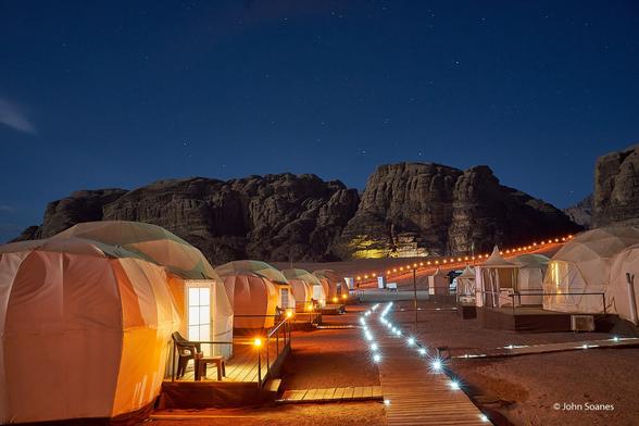 A lit boardwalk winds its way over the tan, desert sand.  The path is flanked with rows of "Martian" tents.  There is a rock formation mid-frame leading to a dark blue night sky filled with stars.