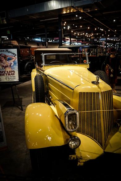 The image features a vintage yellow car prominently displayed in what appears to be a museum or exhibition space. The car has a classic design with a large, vertical grille and rounded fenders, characteristic of early 20th-century automobiles. It has a convertible top and large, round headlights, with a spare tire mounted on the front fender. The car's paint is glossy and well-maintained, reflecting the overhead lights.

In the background, there is a movie poster for "My Lips Betray" featuring an image of a woman, suggesting a historical context. The setting includes other vintage vehicles and exhibits, with a person visible in the background, adding a sense of scale and activity to the scene. The lighting is warm and ambient, enhancing the nostalgic atmosphere of the exhibit.

Provided by @altbot, generated privately and locally using Ovis2-8B