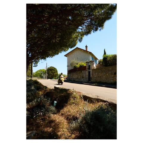 Wide angle image taken at noon on a hot, sunny day in a small village in Provence. The foreground is framed by the branches of a tree and bushes growing on the side of the road. The road diagonally through the frame from the center left to the bottom right. A person on a scooter, blurred by the slow shutter speed, is approaching from the left. A single two story building is visible in the background.