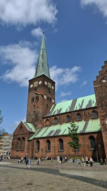 Upright photo. Standing on the main square in Aarhus, Denmark looking at Domkirken, Aarhus Cathedral, completed in the year 1500. The sun is shining, the sky is blue, people are walking and cycling by. The cathederal is 93m long and redbrick with copper-green spires. The 96-metre-tall clock and bell tower rises above everything. Flapping down in high wind from a window way up in the belfry: a giant Pride banner, several metres long. It's to celebrate the annual Aarhus Pride festival with parade today.