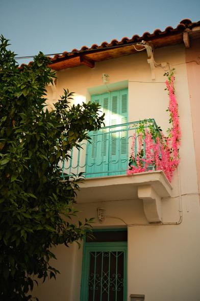 A house in a city with a cyan door. Above the door is a small balcony also with a cyan door. A ray of sunshine is shining on the balcony door. There is also a pink plant on the right side of the balcony (possibly fake). A dark green tree is covering the entire left side of the house.