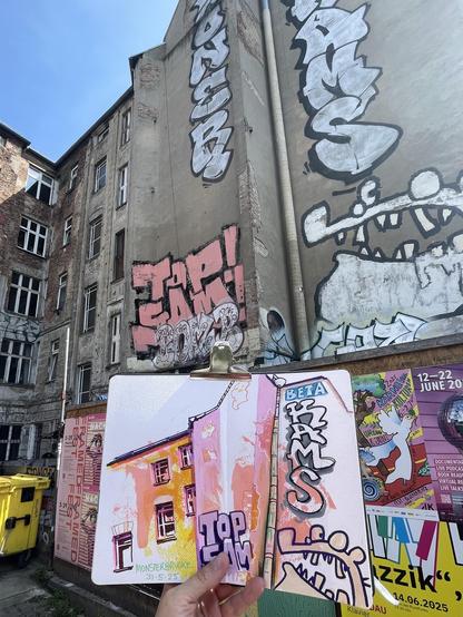 A hand is holding a sketchbook with a sketch of the view behind- a house front with graffiti and windows.