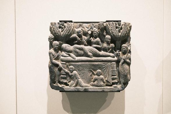 The passing away of the Buddha, Pakistan, Gandhara, 2nd-3rd c., schist. At the age of 80, the Buddha consumes a spoiled meal while traveling. Weakened, he lies down in a grove to die. He gives his monks encouragement one last time and gives instructions on how to deal with his mortal remains. Then the Buddha finally enters Nirvana. The relief depicts monks and laypeople gathering to mourn at the deathbed. Exhibited at the Humboldt Forum in Berlin, 2023.