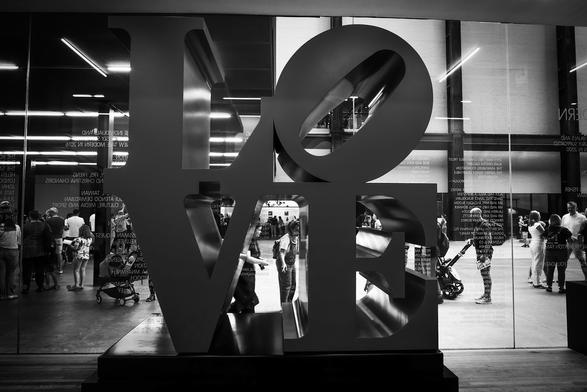 Black and white. Inside the Tate Modern art gallery at Bankside by the River Thames in London, is a large sculpture, perhaps fifteen feet square, of the word "LOVE", with the "LO" on top and the "VE" below, the "O" being slanted at 45 degrees to the right. The original is by Robert Indiana. Through a wide multi panel window behind it visitors to the gallery can be seen.