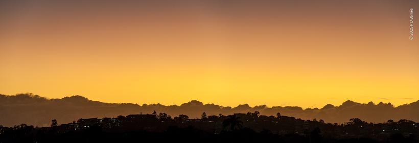 A panoramic sunrise. Below, a gently convex suburban ridgeline lies still dark, while immediately above gently concave bank of clouds makes a second silhouette. Above them  both is a soft, saffron-rice yellow and orange pre-dawn sky. (The joke refers to the vague visual similarity of the image to a doublet lens with a concave element glued to a convex element to reverse the differential bending of different wavelengths of light, causing chromatic aberration)