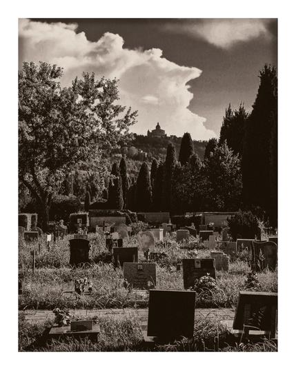 The photo is of cemetery with headstones of various shapes and sizes - with grass and scattered flowers around them. In the background there is a row of manicured cypress trees which helps lead your eye toward a hilltop with a silhouetted church (San Luca) which is in the mid ground of the photo. Above the hill in the background, and takes up roughly the too third of the photo, there are dramatic clouds in the sky.