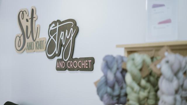 Sign reading “Sit and Knit, Stay and Crochet” above yarn shelves in AriYARN Shop and Studio, Park Ridge, IL.