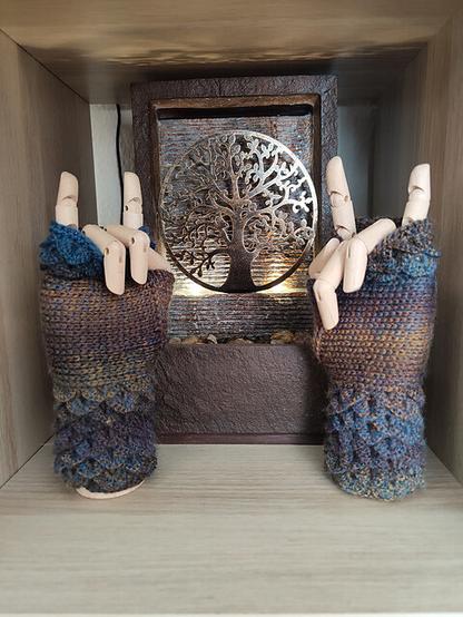 A pair of crocheted mittens with a textured, scale-like pattern is displayed on a light surface. The mittens have an intricate design, resembling dragon scales, and are made in a mix of deep blue, gray, and brown tones. The cuffs are ribbed, providing a snug fit. The yarn used for the mittens has a soft and slightly variegated color effect, adding depth to the scale pattern