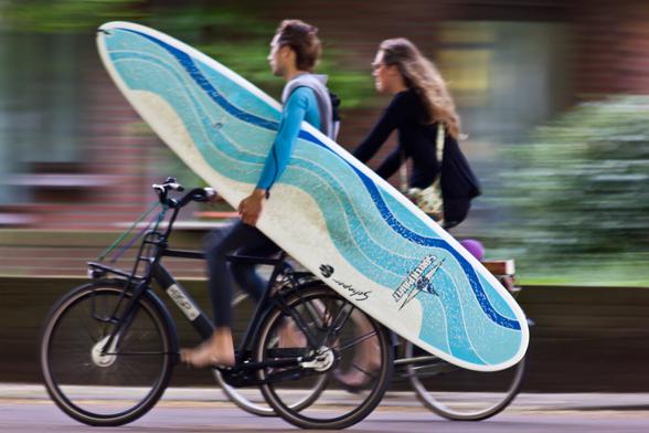 In the middle of the photo two cyclists next to eachother, photographed from the side. In front a young man and behind him a young woman. The man is cycling barefoot and has a large white surfboard (printed with blue waves) under his left arm. Only his right hand is on the handlebars. Due to the speed and movement some parts are blurred, such as the feet and the background.