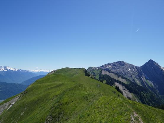 View back to the grassy ridge with at the end Le Grand Roc summit. Some rocky mountains on the right and snow capped mountains on the left.