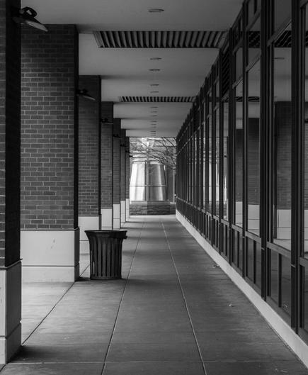 Image of a sidewalk covered by part of a building alongside it. The shot is taken more-or-less straight-on toward the end of the corrdior, with the image attempting to frame the rectangular end of the walkway dead-center, with perspective lines leading toward it.