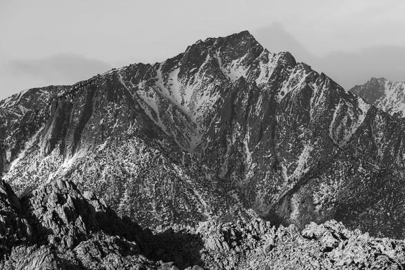 A dramatic black and white image of a rugged mountain range, featuring steep rocky peaks and patches of snow. The foreground includes jagged rock formations, while the background shows a towering peak against a cloudy sky.