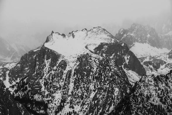 Black and white image of a rugged mountain landscape, featuring snow-covered peaks and rocky slopes, partially obscured by mist. Dense patches of trees are visible on the lower slopes.