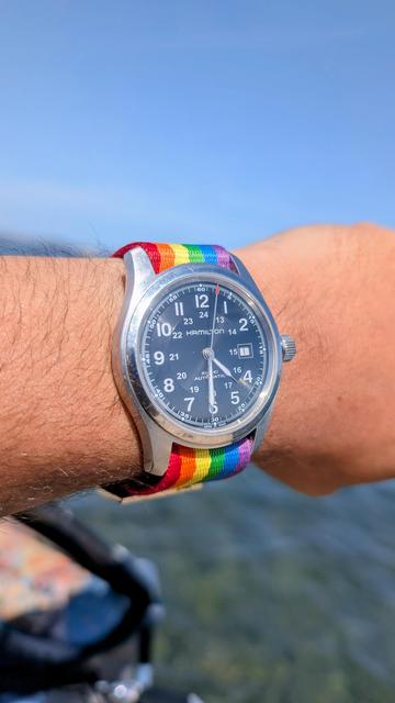 Photo de mon poignet, et de ma montre avec un bracelet arc en ciel, sur fond de mer floue et de ciel bleu.
