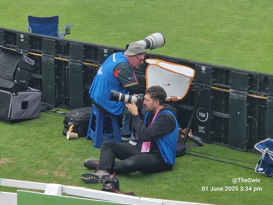 The image depicts two photographers on a grassy field, likely at a sports event. The photographer on the left is seated on a blue stool, wearing a blue vest with a pink lanyard, and is adjusting a camera with a large telephoto lens. The photographer on the right is sitting on the ground, also wearing a blue vest, and is focused on taking a photograph with a camera. Both photographers have professional equipment, including large camera bags and a reflector. The background features a black barrier with the logo "TGI" and a blue folding chair. The date and time, "01 June 2025 3:34 pm," are visible in the bottom right corner, along with the watermark "[@]TheSwiv."

Provided by @altbot, generated privately and locally using Ovis2-8B