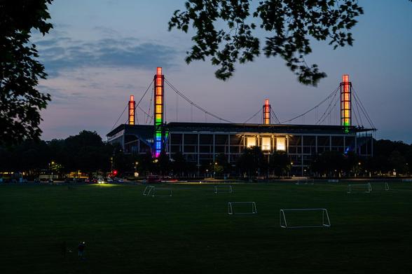 Das Fussballstadion in Köln-Müngersdorf am Abend. Die 4 Masten sind im pridemonth in Regenbogenfarben beleuchtet.links und oben sind Äste von Bäumen zu erkennen. Auf den Vorwiesen des Stadions sind Fussballtore zu erkennen.
