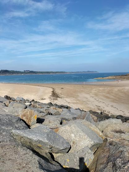 Photographie d’une plage bretonne à marée basse, vue depuis un amas de rochers gris. Au loin, une mer bleu profond sous un ciel clair légèrement strié de nuages. Quelques promeneurs minuscules profitent du calme.
