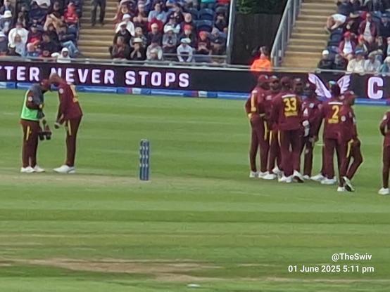The image depicts a cricket match scene. In the foreground, a group of cricket players wearing maroon uniforms with yellow accents are gathered, likely discussing strategy or celebrating a moment. The player with the number 33 is visible in the center of the group. To the left, two players in maroon uniforms are engaged in a conversation, with one player wearing a green vest, possibly a coach or support staff. The pitch is visible with stumps in the center, and the outfield is green and well-maintained. In the background, spectators are seated in the stands, and a large electronic billboard displays the text "BETTER NEVER STOPS." The date and time "01 June 2025 5:11 pm" are visible in the bottom right corner, along with the watermark "[@]TheSwiv."

Provided by @altbot,