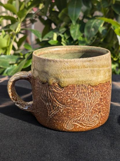 Front view of a small, brown and green mug. The form is roughly as wide as it is tall, with a rounded handle. An abstracted fire/water motif is carved on the bottom 2/3 of the surface, enveloped at the top by a shiny and ever so slightly drippy moss green glaze.