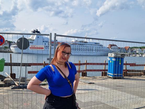 Das Bild zeigt eine Frau mit roten Haaren und Brille, die vor einem Hintergrund mit einem großen Kreuzfahrtschiff am Hafen steht. Sie trägt ein blaues, kurzärmeliges Oberteil mit Ausschnitten und schwarzem Rock. Ihre Hände sind auf dem Hüftbereich abgestützt, und sie lächelt in die Kamera. Im Hintergrund ist ein großes weißes Kreuzfahrtschiff zu sehen, das am Hafen liegt. Es gibt auch ein Schild mit der Aufschrift "MAC Meet & Greet" und ein weiteres Schild mit einem "No Parking" Symbol. Der Himmel ist teilweise bewölkt, und im Hintergrund sind Häuser am Ufer zu erkennen. Es gibt auch ein blaues Müllbehältnis und ein Metallzaun im Vordergrund.