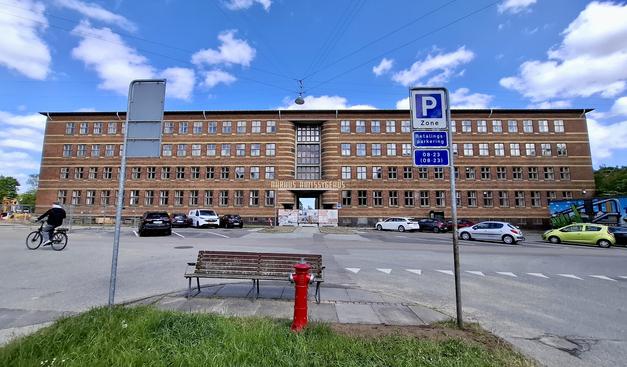 Looking at the main building at the entrance to the vast former county hospital, near mid-city Aarhus, Denmark. Aarhus Amtssygehus (County Hospital) was used from 1883 to 2018. The main building we’re looking at was built in the 1880s. It is very long and five storeys high with rows of windows. It has characteristic red brickwork with single rows of yellow bricks, like ribbons, under the row of windows of each storey. Some cars are parked outside, a cyclist is just passing, there’s a bench and signage and a red fire hydrant on a patch of grass just in front of us. The sky is deep blue and dotted with white clouds.