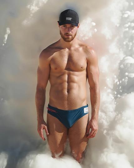 A muscular white guy wearing speedos is standing in front of a big wave of bubbles