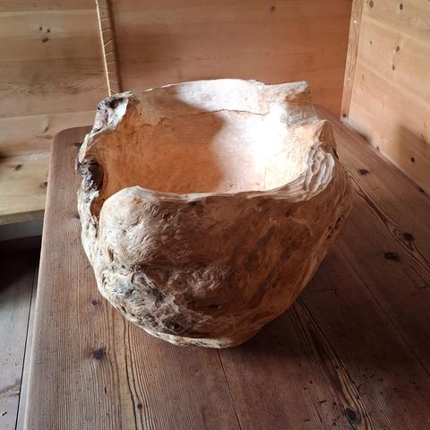 A large, organically shaped natural woodform rests on a larch wood table. The room features warm-toned, knotty arolla pine paneling and built-in bench seating. The object appears to be carved from a single, solid piece of wood, with flowing curves and hollowed areas that resemble an abstract bowl or natural seat. The surface showcases a mix of smooth, pale sections and rough, textured bark-like areas, emphasizing its raw and sculptural qualities.