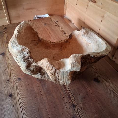 A large, organically shaped natural woodform rests on a larch wood table. The room features warm-toned, knotty arolla pine paneling and built-in bench seating. The object appears to be carved from a single, solid piece of wood, with flowing curves and hollowed areas that resemble an abstract bowl or natural seat. The surface showcases a mix of smooth, pale sections and rough, textured bark-like areas, emphasizing its raw and sculptural qualities.
