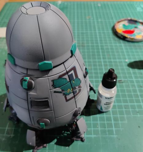 A grey Overlord-C DropShip with a half-hull tall Clan Jade Falcon emblem painted on its side. The miniature is coated with gloss varnish which doesn't really show in the photo at all. The varnish bottle is next to the mini for scale, on my green well-worn cutting mat.