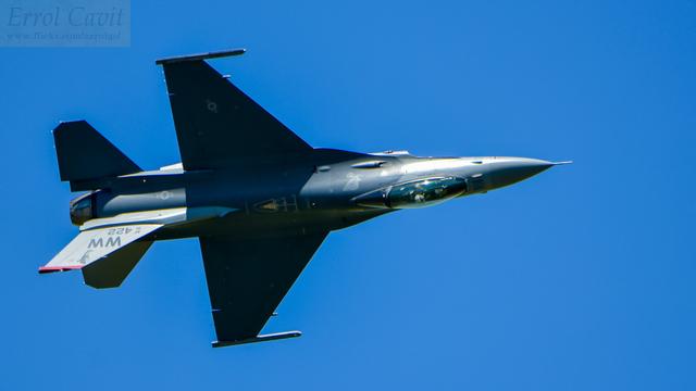 Fighter jet rolling as it zooms past, just starting to go inverted. The fuselage and fin are lit by the strong sun, while the wings and horizontal stabilisers are in shadow.