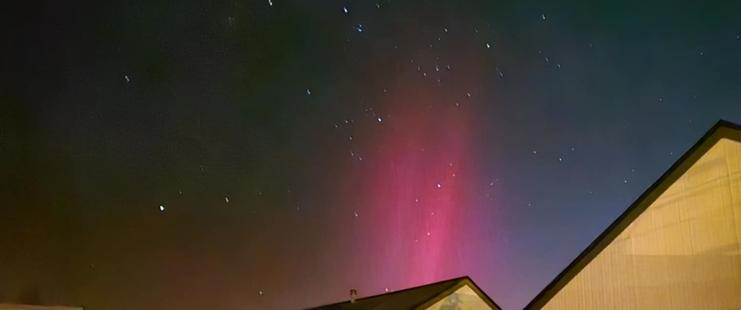 Photo of the night sky with a lot of stars showing.  There are the tops of some neighbouring houses in the foreground, particularly on the right.  The aurora is shining up from the bottom of the photo just off center left.  It is mostly 2 beams of magenta/red pointing upwards and angled slightly right.