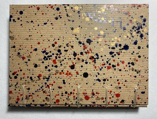 La couverture d'un carnet en papier kraft moucheté de projections d'encre :  noir, rouge, doré. On voit les passage des deux ficelles de la reliure qui partent vers le dos du livre.