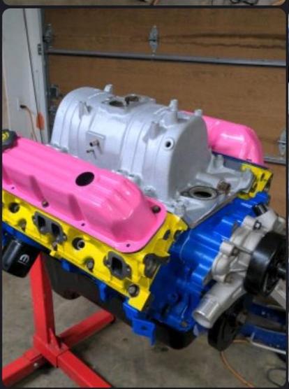 A Dodge 5.2 liter V8 engine, on a stand in a garage. The major engine components have been painted to resemble the pan flag colors (pink valve covers, yellow heads, blue block). The other components are either silver or black.
