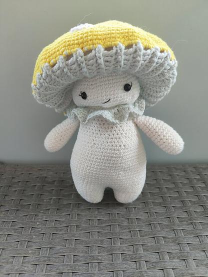 A crocheted plush toy resembling a cute mushroom character. The toy has a soft, rounded body in muted white with two small arms and legs. It features a smiling face with black eyes and a stitched mouth. On its head is a large, bright yellow mushroom cap with light gray frilly edges underneath, resembling gills. The toy sits on a woven gray surface with a plain light background.