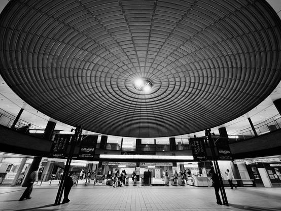The lobby of an underground station in Yokohama is dominated by a huge tiled disk decorating the ceiling. The dark textured tiles form concentric circles. There is a pattern of spokes leading to two bright lights at the centre. At ground level people are hanging around, leaning on pillars. There are shop fronts in the background. The striking design looks like a UFO descending.