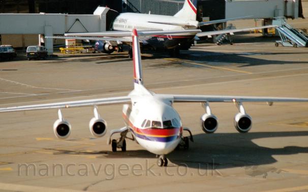 directly towards the camera.
The plane is white, with a red and purple stripe with yellow piping in the middle running along the body, and a red tail with a purple stripe with yellow piping at the bottom.
It's nose wheel is turned to it's left, as it turns to the right of the frame.
In the background, a grey and dark blue, twin engined jet airliner facing away from the camera, has an airbridge connected to it's forward fuselage, the other end of the bridge connected to  a grey building.