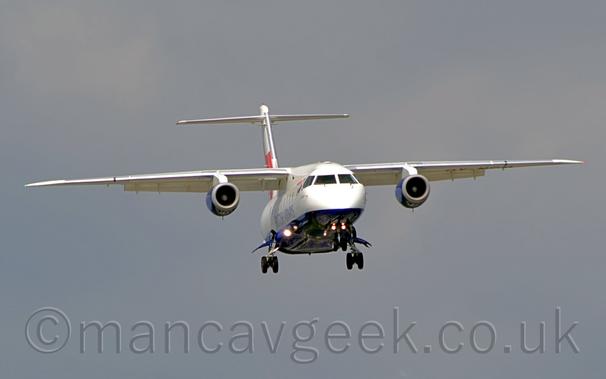 Front view of a high-winged, twin engined jet airliner flying almost head-on towards the right of the camera at a very low altitude, with undercarriage lowered, and flaps extended from the rear of the wings, suggesting it is about to land.
The plane is mostly white, with a blue belly, and blue "British Airways" titles  on the side of the lower fuselage.
The engine pods mounted under the wings are the same dark blue, while the tail is white and red.
Flat grey sky fills the rest of the frame.
