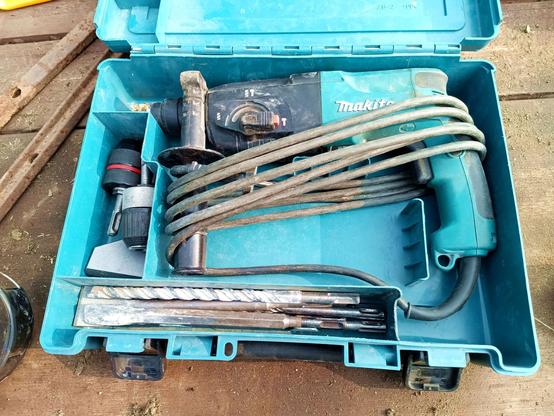 A Makita hammer drill in its teal coloured case, a sturdy box with compartments. The main compartment holds the machine with the cable wrapped around both handles. The two support baffles are designed with a gap for the cable to pass through. The lower compartment has a selection of well used masonry drill bits and chisels, the left one two drill chuks with SDS fittings. Everything is a bit dusty but not dirty.