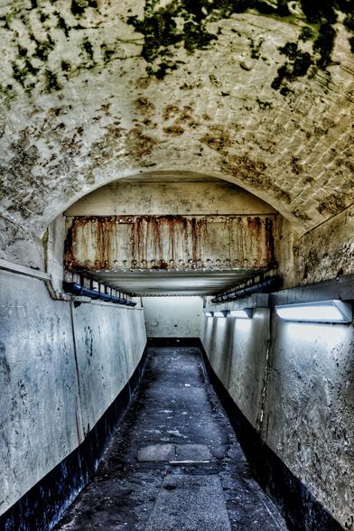 A dimly lit, narrow, arched tunnel or passageway stretches into the distance. The ceiling and upper walls are rough, stained concrete with patches of dark mould or discolouration. A rusty metal beam spans across the width of the tunnel near the top, with rust streaks running down the wall beneath it.
Along the lower parts of both side walls are angled concrete surfaces, beneath which are dark, possibly wet, areas. Modern, rectangular strip lights are mounted on the lower part of the right-hand wall, casting a bright, cool light onto the scene. The floor is a dark, uneven surface. The far end of the tunnel appears brighter, suggesting an exit or another light source. The overall impression is of a damp, somewhat derelict, underground structure.