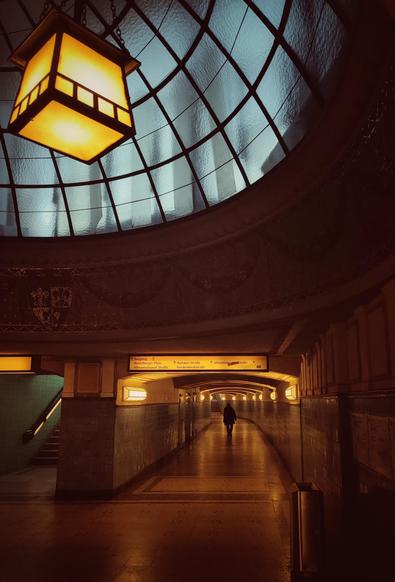 Blick in einen Fußgängertunnel im U-Bahnhof Heidelberger Platz. Die Farben sind warm und gelblich/orange, von hinten ist die Silhouette einer Person zu erkennen, die durch den Tunnel läuft. Links oben sieht man eine Kuppel aus Glasfenstern, durch die aber nur künstliches Licht fällt. Ganz oben in der Kuppel hängt eine große eiserne Lampe mit gelblichem Licht.