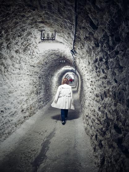 The portrait-orientated image depicts a woman viewed from behind, walking down a long, dimly lit, arched tunnel. The tunnel's walls and ceiling are rough and textured, appearing to be made of natural rock, with visible white and off-white tones. Overhead, there are periodic light fixtures providing illumination, with wires running along the ceiling.
The woman is wearing a light-colored coat that reaches below her hips, and dark trousers. Her hair is reddish-blonde. Further down the tunnel, in the distance, a group of other people can be seen, appearing smaller due to perspective, suggesting the tunnel is quite long. The floor of the tunnel is a relatively flat, concrete-like surface, with some visible darker patches.