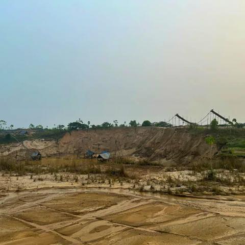 A barren, scarred landscape from suction gold mining in Balata, Peru, showing deep excavated pits, sandy tailings piles, and muddy water pools where Amazon rainforest once stood. Mining equipment and structures are visible in the background, with a thin line of remaining forest on the horizon.