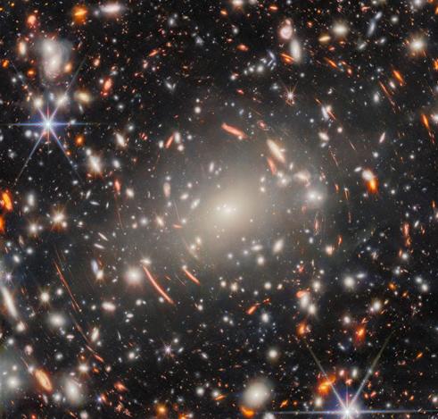 A field of galaxies in space, dominated by an enormous, bright-white elliptical galaxy that is the core of a massive galaxy cluster. Many other elliptical galaxies can be seen around it. Also around it are short, curved, glowing red lines, which are images of distant background galaxies magnified and warped by gravitational lensing. A couple of foreground stars appear large and bright with long spikes around them.]
